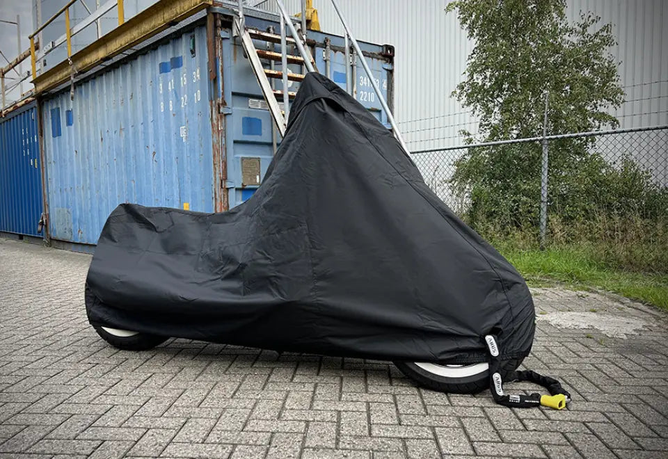 Sort motorcykelovertræk med lås foran en blå container.